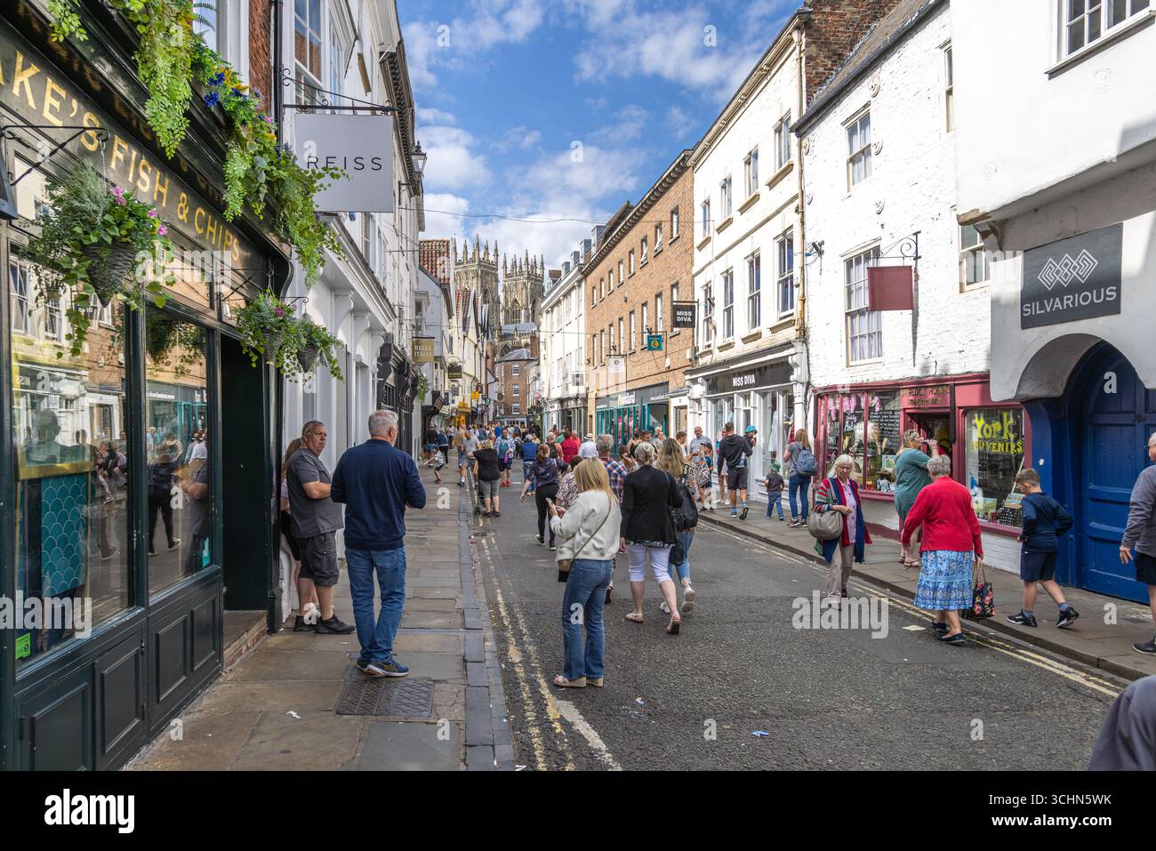 Turisti e gente del posto in una strada trafficata di York, in Inghilterra, con edifici storici, negozi e guglie della cattedrale di York che si innalzano sullo sfondo. Foto Stock