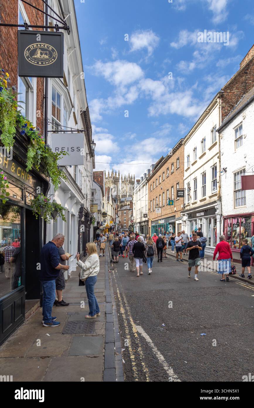 Turisti e gente del posto in una strada trafficata di York, in Inghilterra, con edifici storici, negozi e guglie della cattedrale di York che si innalzano sullo sfondo. Foto Stock