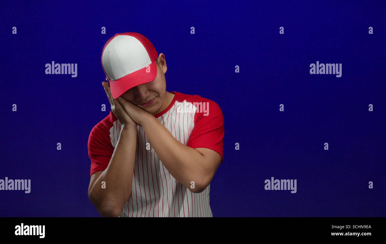 Giovane cinese in uniforme da baseball con strisce rosse e bianche che si posa sonnamente su sfondo blu isolato, suggerendo un atleta stanco Foto Stock