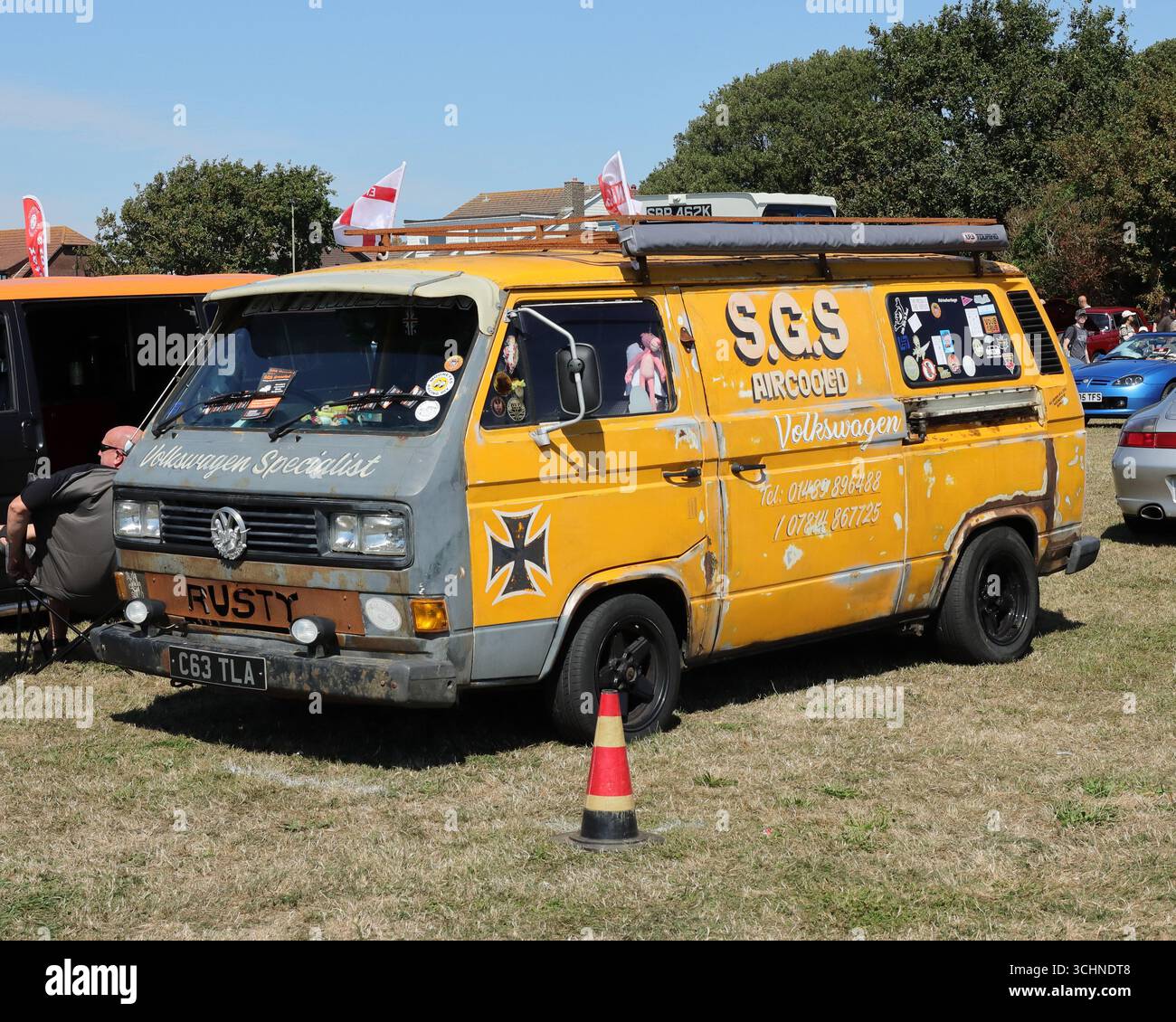 Questa foto mostra un furgone Volkswagen T3 giallo intemprato, decorato con adesivi e un cartello "RUSTY". Ha un portapacchi ed è parcheggiato su un campo di erba secca. Un piccolo cono stradale si trova di fronte alla ruota e altri veicoli e persone sono visibili sullo sfondo sotto un cielo luminoso e soleggiato. Illustrativo, editoriale: Lo Stokes Bay Car Rally a Gosport, Hampshire, Inghilterra. 25 agosto 2025. Foto Stock