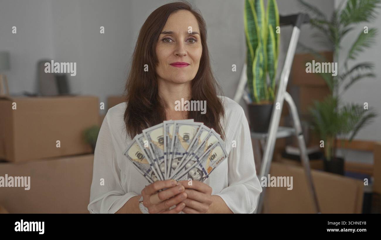 Donna che tiene dollari al chiuso in una nuova casa circondata da scatole e piante in movimento, che celebra il successo finanziario con la nostra valuta, sorridendo felicemente in California Foto Stock