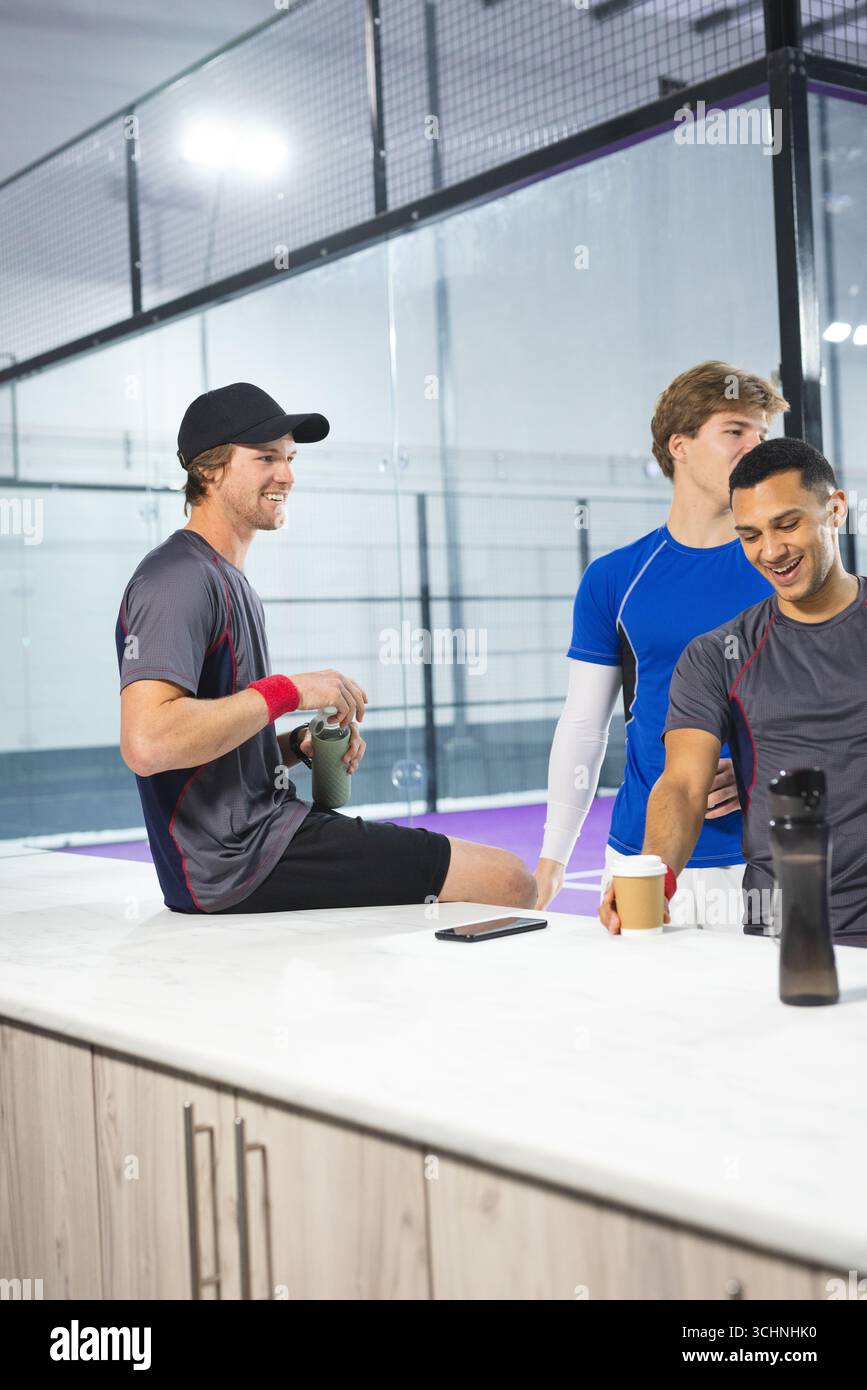 Diversi amici maschili in abbigliamento sportivo appoggiati al bancone e sorseggiano bevande presso il campo di vetro del padel Foto Stock