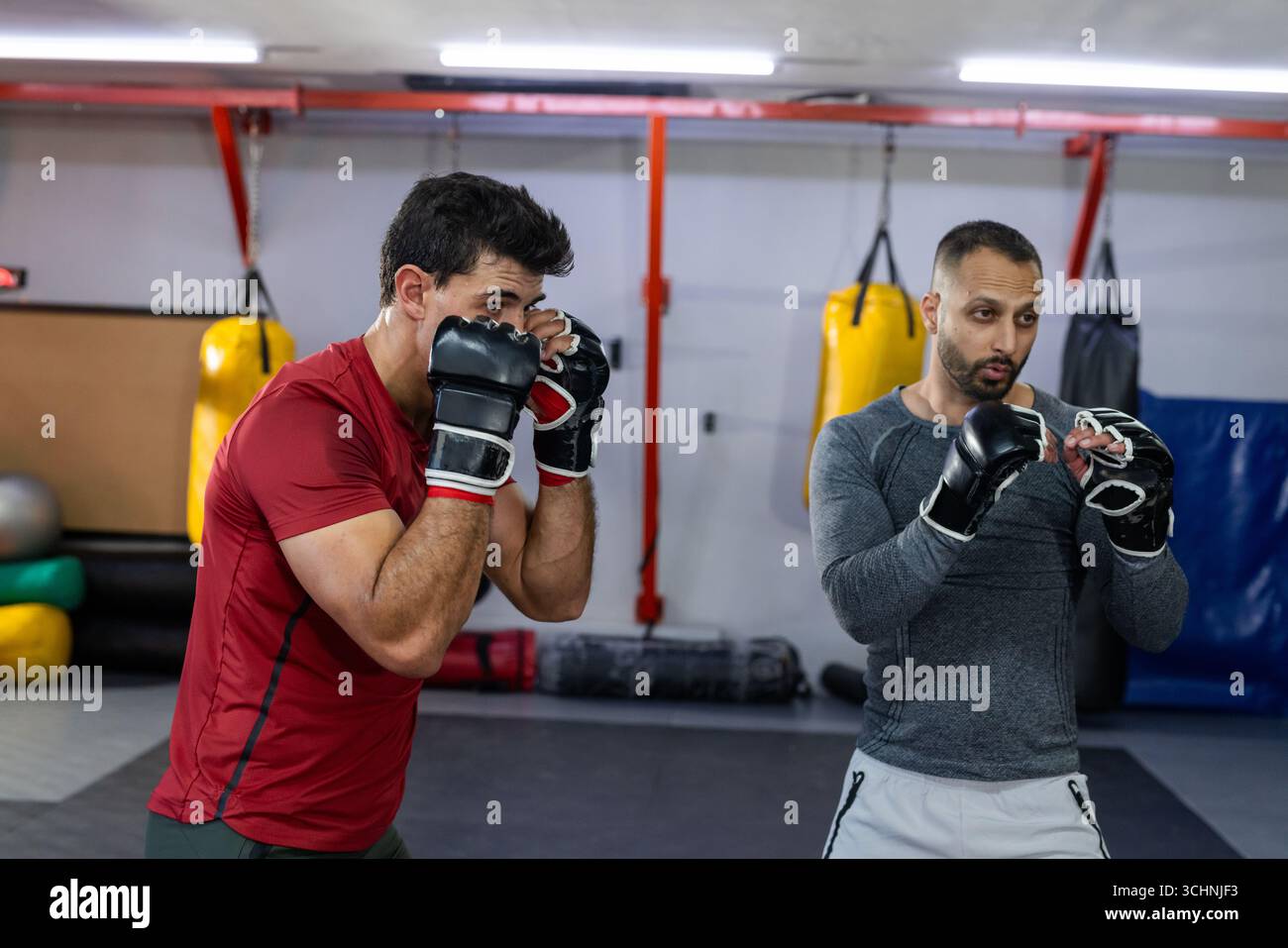 Allenatore maschile variegato, tirocinante che pratica colpi sul tappeto imbottito nella palestra da combattimento con guanti mma Foto Stock