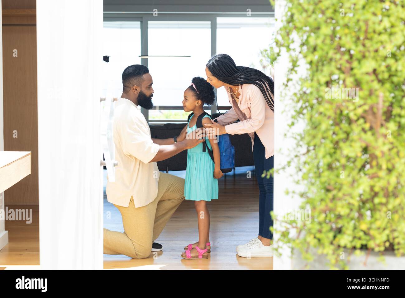 I genitori aiutano la figlia con lo zaino, si preparano per la scuola a casa, sorridono calorosamente Foto Stock