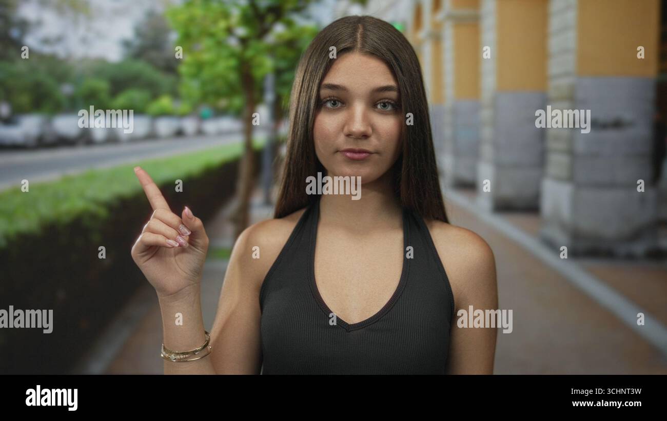 Una ragazza in cima nera punta un dito rialzato lungo una strada urbana rigogliosa fiancheggiata da auto parcheggiate ed edifici in pietra; curiosità. Foto Stock