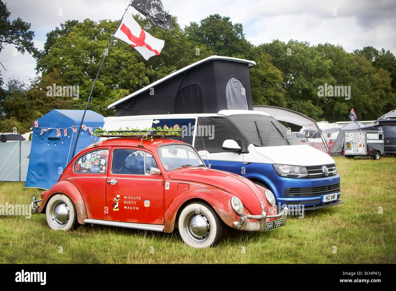 Camper vintage rosso Volkswagen Beetle e moderno VW, parcheggiati in un campeggio con tende e alberi sullo sfondo Foto Stock