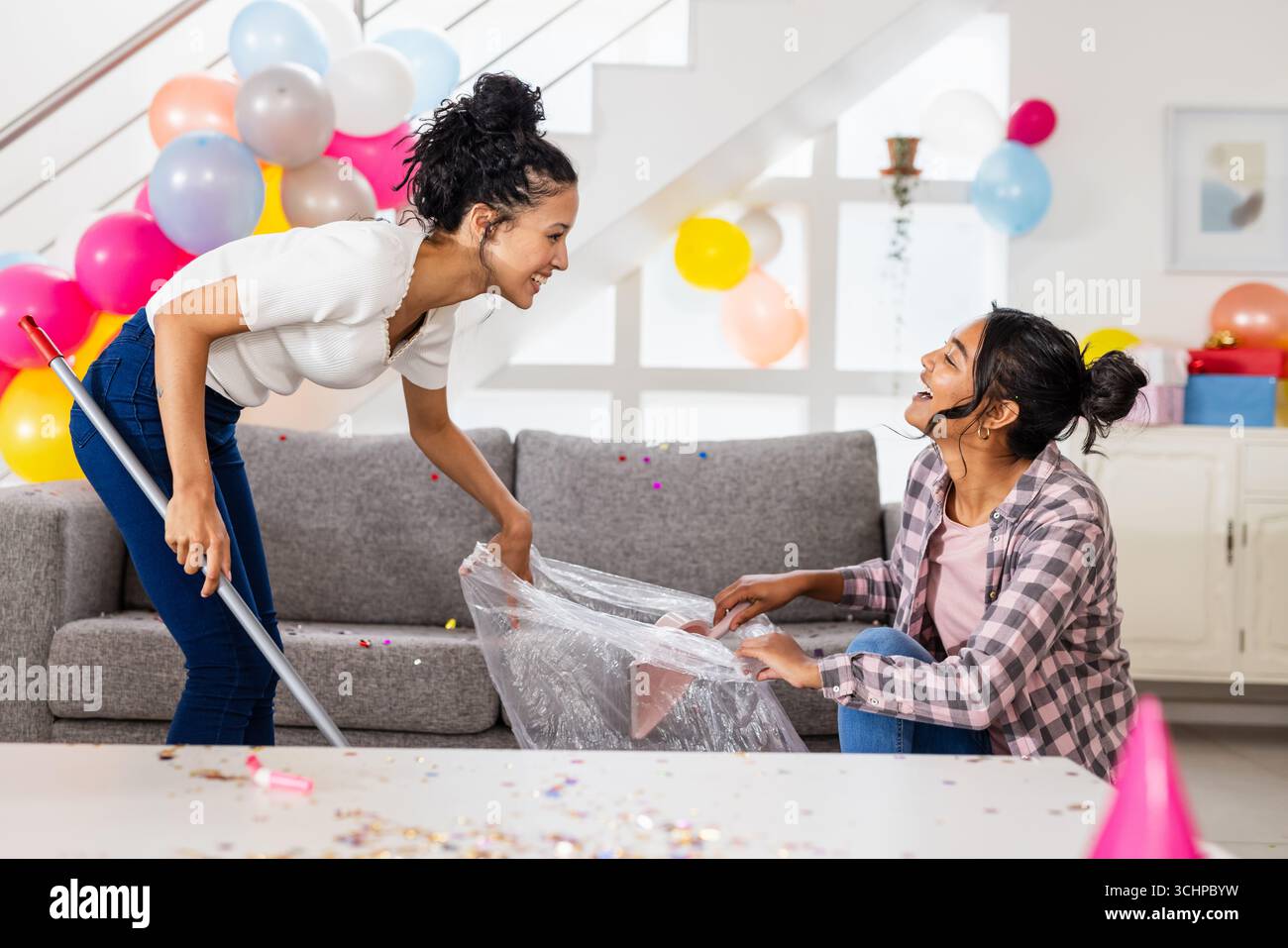 Diverse amiche si ripuliscono dopo la festa a casa, sorridono e si divertono al lavoro di squadra Foto Stock