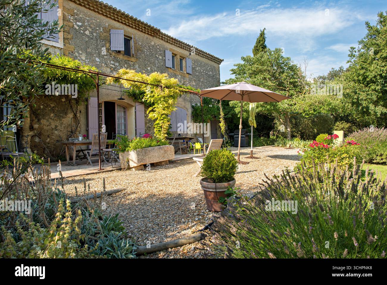 Villa Patio del XVII secolo con ghiaia e lavanda Saint Martin De Crau Francia // SAINT-MARTIN-DE-CRAU, Francia — Un patio di ghiaia si trova di fronte a una villa del XVII secolo in Provence les Alpilles, Francia. La villa presenta una costruzione in pietra e persiane tradizionali. L'area del patio include posti a sedere all'aperto, un barbecue e un grande ombrello, suggerendo uno spazio progettato per il relax e la cena all'aperto. La villa è circondata da una vegetazione lussureggiante, tra cui ulivi e cespugli di lavanda, caratteristica del paesaggio provenzale. La scena cattura il fascino rustico e l'atmosfera serena tipica Foto Stock