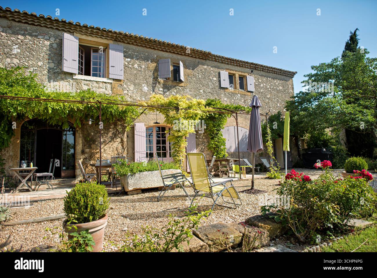 Casa in Provenza Francia Saint Martin De Crau // SAINT-MARTIN-DE-CRAU, Francia — Una casa in pietra con tetto piastrellato e persiane di colore chiaro si vede nella regione francese Provence les Alpilles. L'esterno presenta una grande porta ad arco con porte in vetro e diverse finestre con persiane aperte. Di fronte alla casa, i sentieri di ghiaia conducono ad aree salotto all'aperto con tavoli e sedie. Il verde, tra cui vigne arrampicate e cespugli di rose, circonda la proprietà, caratteristica del paesaggio provenzale. Foto Stock