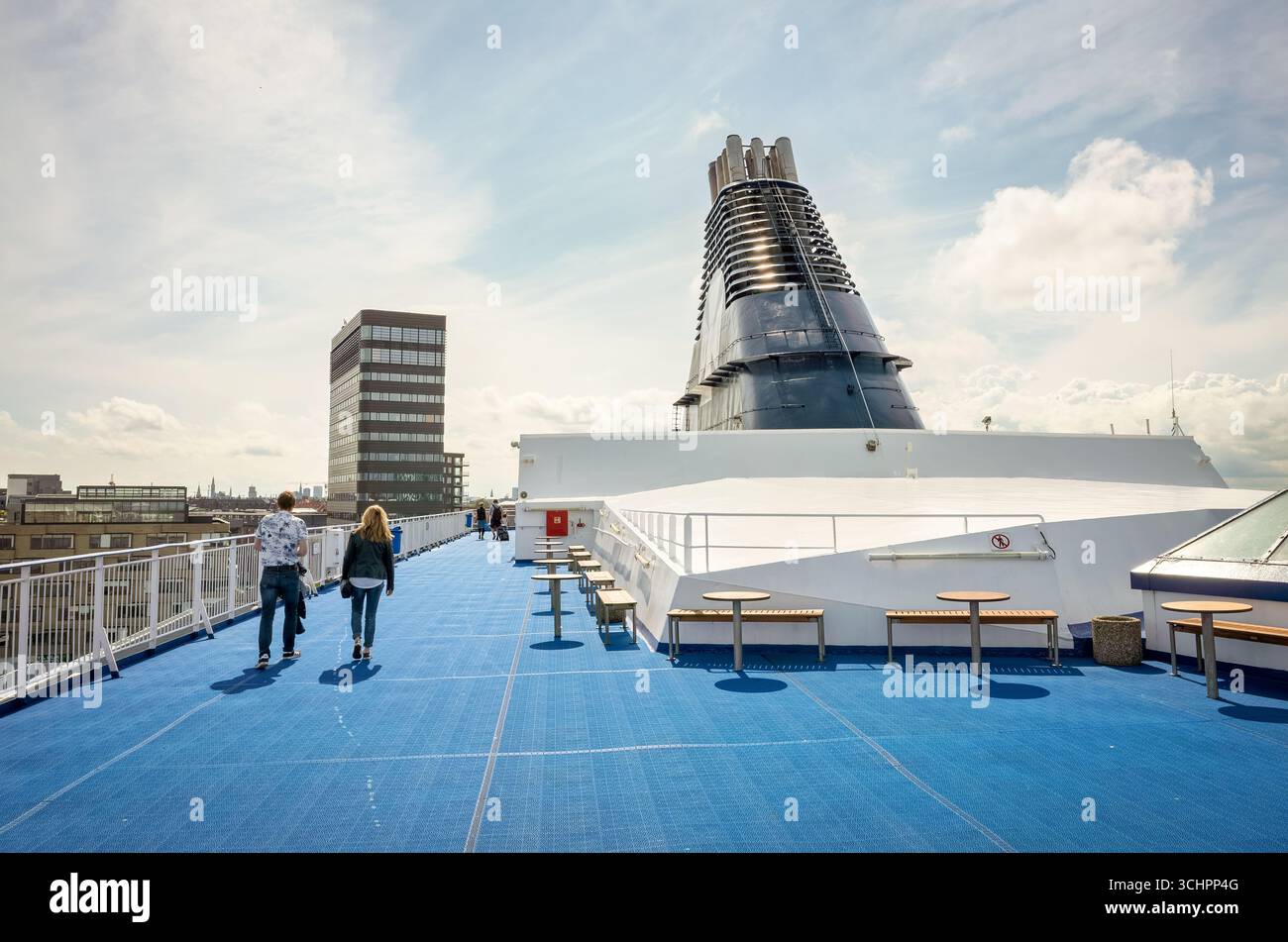 DFDS Ferry Deck Christianshavn Denmark // CHRISTIANSHAVN, Danimarca - Un ponte DFDS Ferry a Christianshavn, Danimarca, con pavimento blu, posti a sedere all'aperto e una vista dell'architettura moderna e dell'imbuto della nave. Due persone camminano lungo il ponte, dotato di tavoli e panchine per i passeggeri. Il servizio di traghetti DFDS gestisce rotte che collegano la Danimarca con altri paesi, tra cui Norvegia e Regno Unito. L'azienda è una delle più grandi società di trasporti e logistica integrate del Nord Europa. Foto Stock