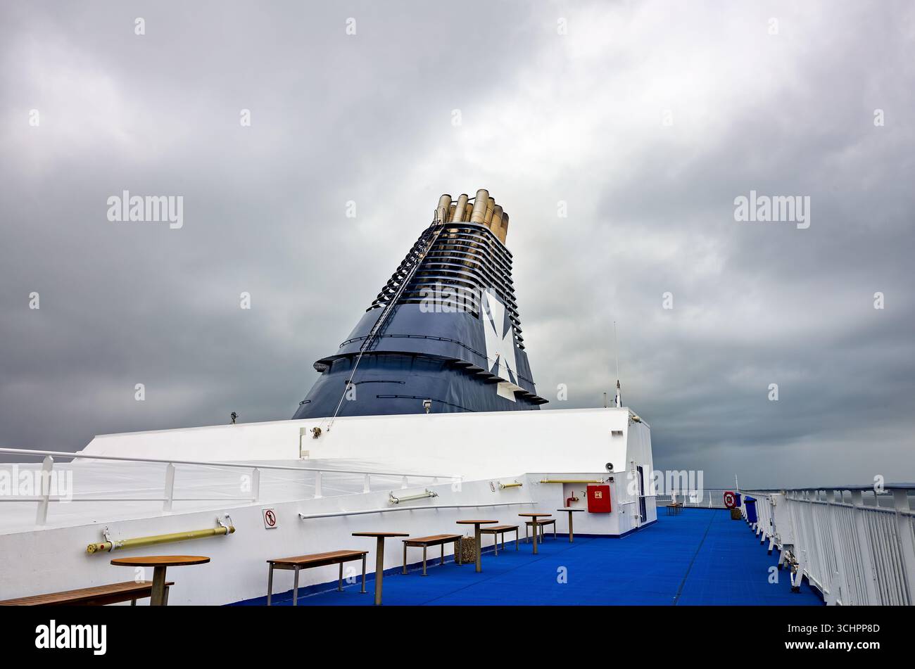 DFDS Ferry Smokestack a Øresund vicino a Rydebäck Svezia // RYDEBÄCK, Svezia — la ciminiera di un traghetto DFDS è visibile contro un cielo nuvoloso, con lo stretto di Öresund sullo sfondo. Il traghetto presenta un grande emblema bianco della croce maltese sulla sua sovrastruttura blu scuro. Il ponte del traghetto è dotato di moquette blu, ringhiere bianche e diverse panche e tavoli in legno. DFDS è una compagnia di traghetti danese-norvegese che opera su rotte attraverso l'Europa settentrionale, compresi i servizi nel Mar Baltico e nella Manica. Le rotte della compagnia collegano diversi paesi, come Danimarca, Norvegia, Swe Foto Stock