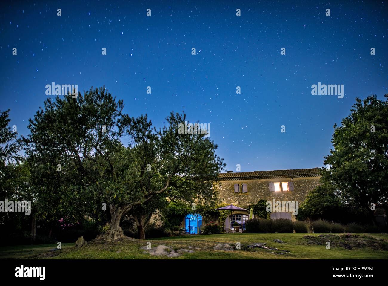 Stelle sopra Villa Saint Martin de Crau del XVII secolo Francia // SAINT-MARTIN-DE-CRAU, Francia - Un cielo notturno limpido pieno di stelle è visibile sopra una villa del XVII secolo in Provence les Alpilles, Francia. La villa, costruita in pietra, presenta persiane tradizionali e un tetto piastrellato. Il paesaggio circostante comprende alberi maturi e terreni curati, creando un ambiente rurale sereno. L'immagine cattura la bellezza tranquilla della campagna francese sotto una vasta distesa celeste. Foto Stock