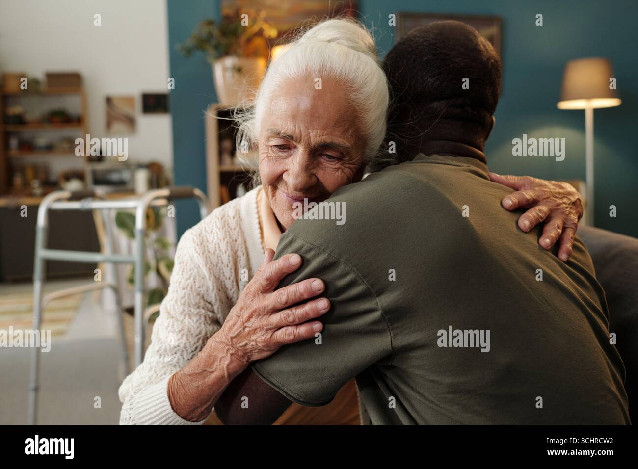 Donna caucasica anziana che abbraccia l'uomo nero in salotto, mostrando affetto e calore, donna anziana sorridente con gli occhi chiusi, camminatore visibile sullo sfondo Foto Stock