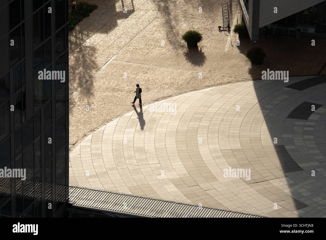 Una persona solitaria cammina attraverso una piazza urbana illuminata dal sole, gettando una lunga ombra sul terreno con motivi geometrici. La scena cattura le linee architettoniche Foto Stock