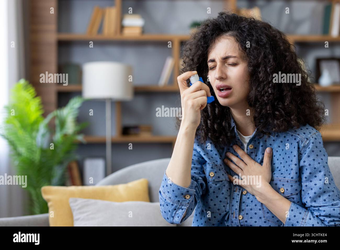 Giovane donna seduta su un divano a casa, che usa un inalatore durante un attacco d'asma, lotta per respirare, tiene il petto in disagio e affronta una crisi respiratoria Foto Stock