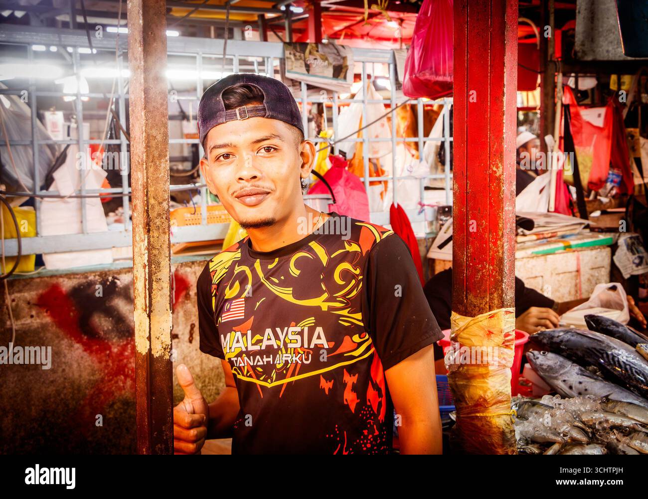 Un venditore di pesce sorridente in un vivace Chow Kit del mercato malese, che indossa un berretto e una camicia vibrante, vicino a prodotti e prodotti colorati Foto Stock