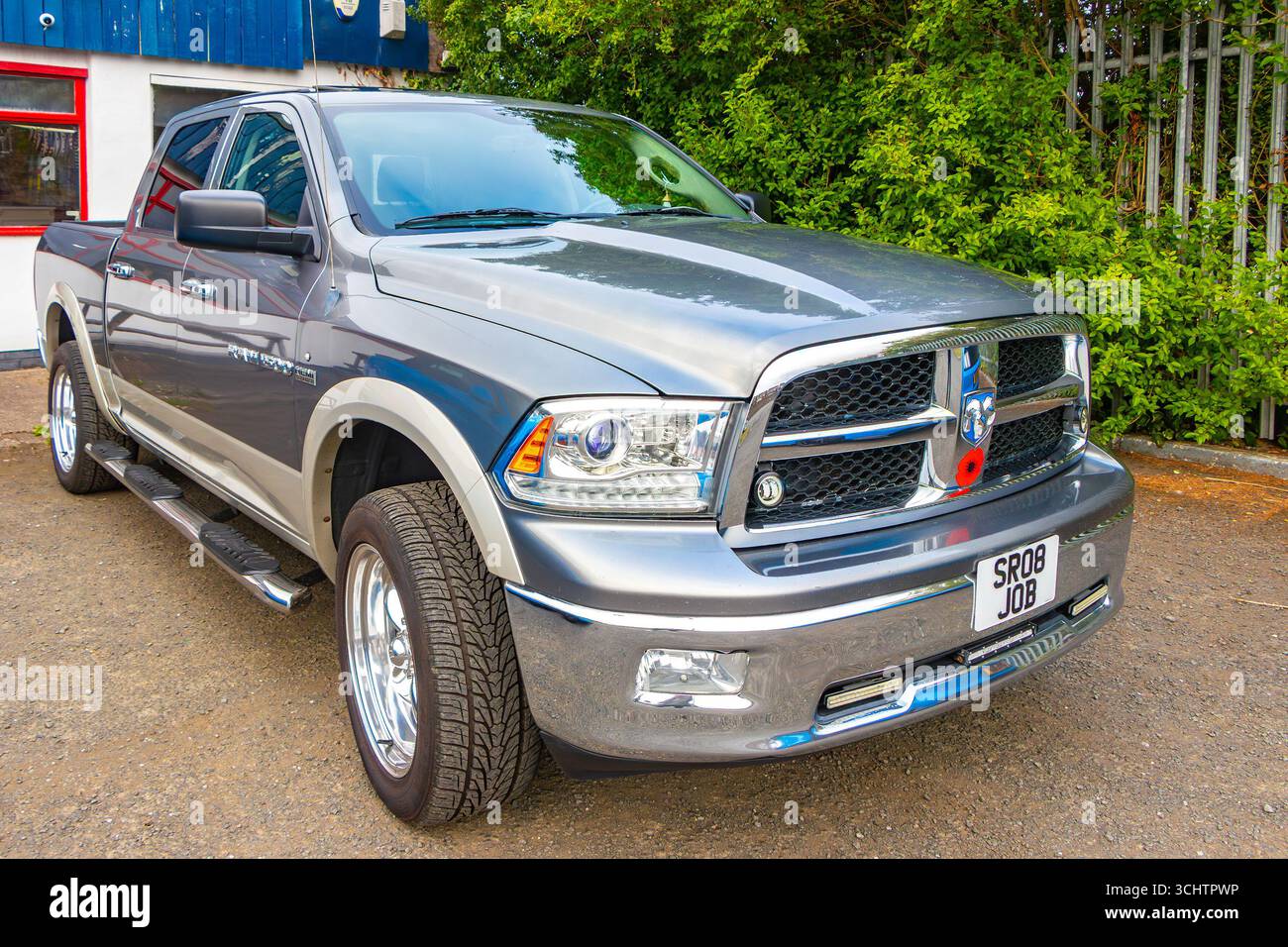 Pick-up Silver Dodge RAM parcheggiato in un sacco di vegetazione intorno e un edificio industriale blu sullo sfondo Foto Stock
