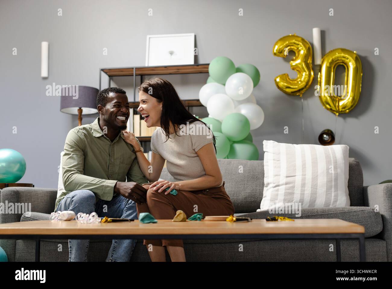 Coppie diverse che ridono insieme sul divano per festeggiare il 30° compleanno con palloncini Foto Stock
