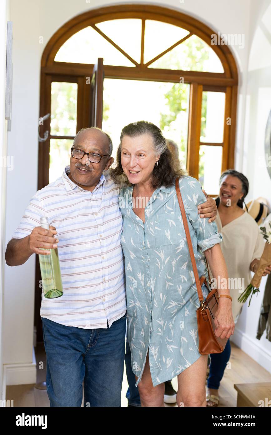 Coppia senior che arriva a casa, tenendo in mano il vino e sorridendo calorosamente con amici anziani diversi Foto Stock