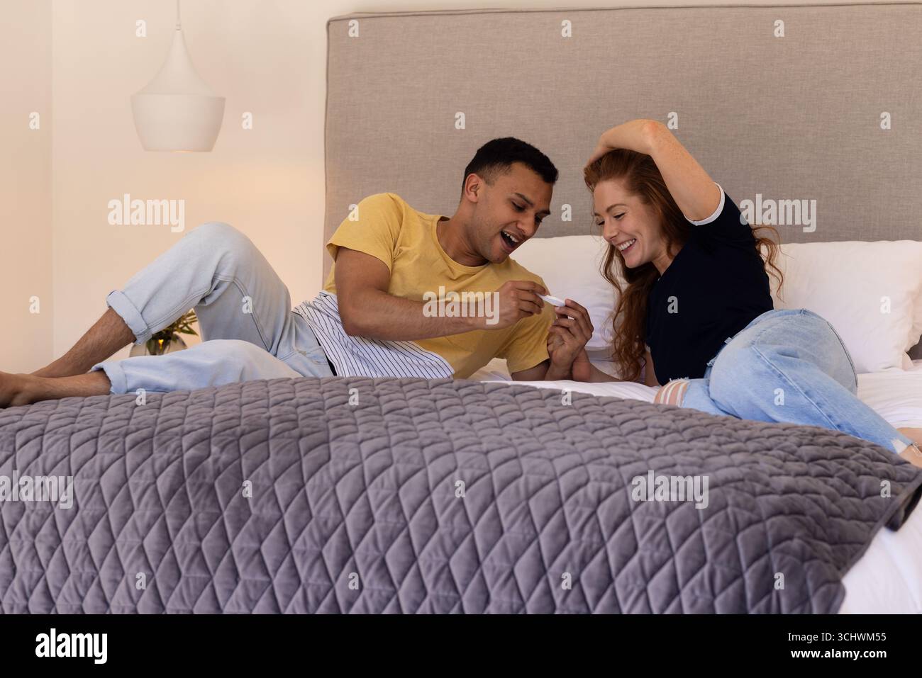 Coppia giovane e diversificata che guarda al test di gravidanza positivo insieme su un letto accogliente a casa Foto Stock