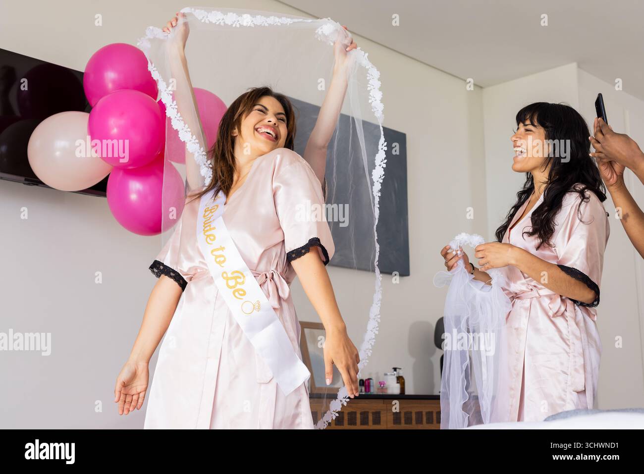 La sposa festeggia con gli amici in vestaglia, tenendo il velo e sorridendo con gioia Foto Stock