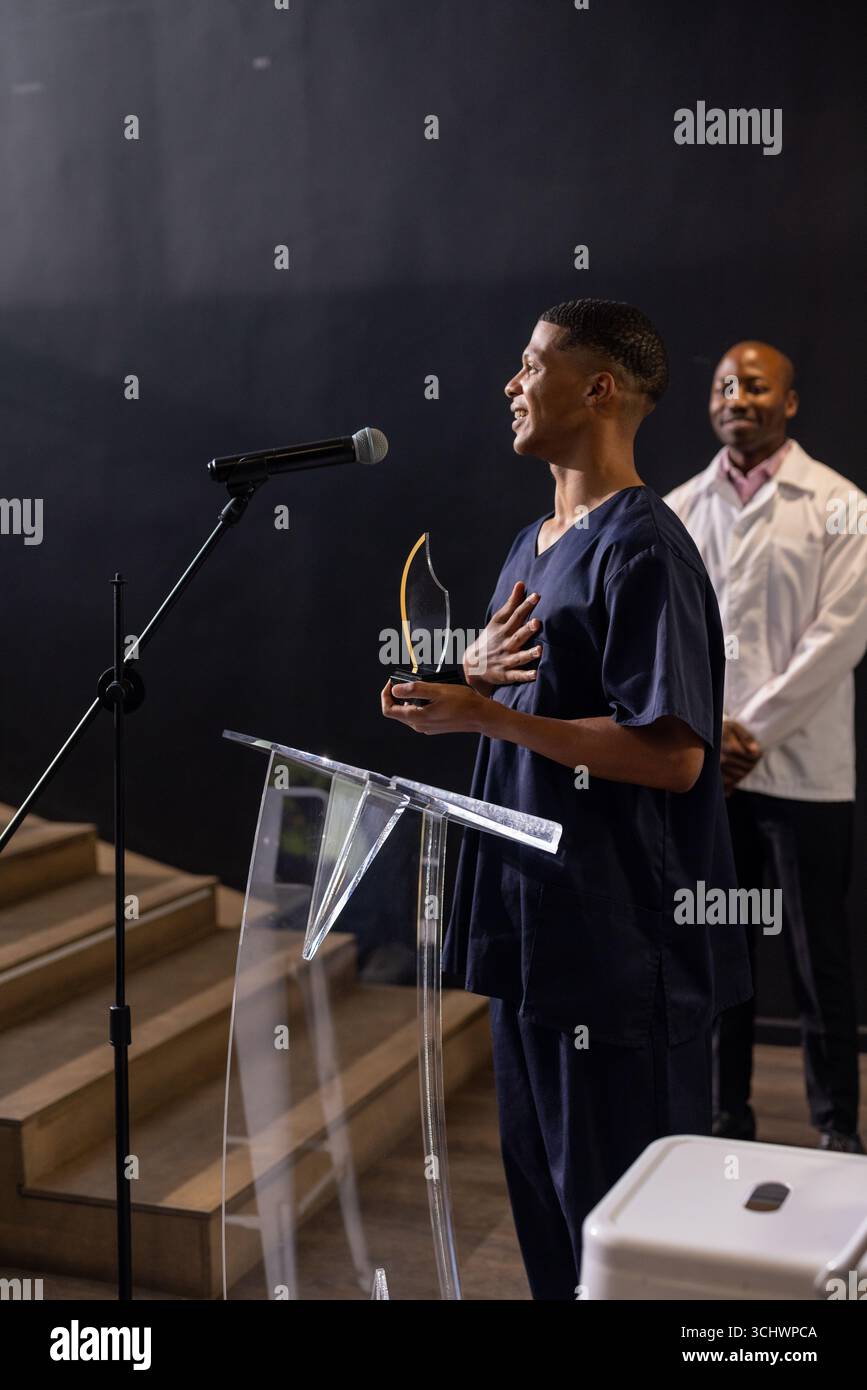 Uomo in scrub che tiene il premio, parla sul podio, collega sorride sullo sfondo Foto Stock