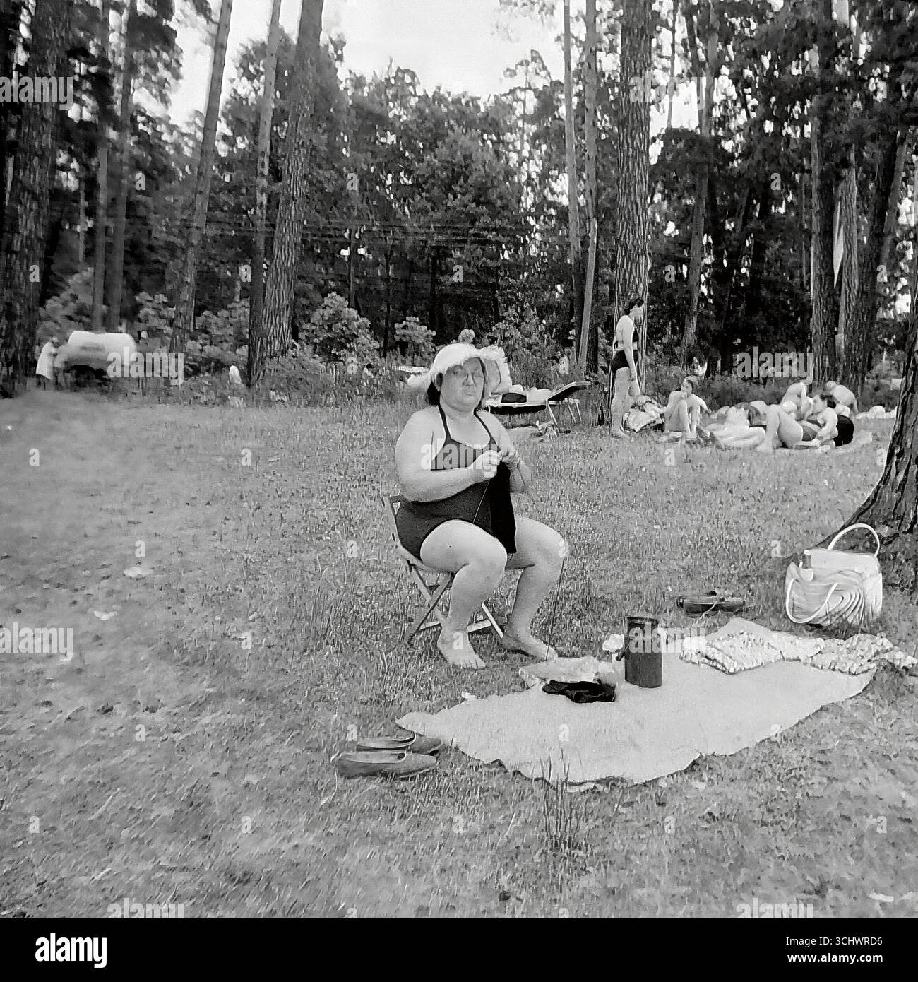 Una fotografia d'archivio in bianco e nero scattata in URSS intorno al 1965. Una donna anziana con costume da bagno e cappello da sole siede su una sedia pieghevole in una pineta e si intreccia durante una ricreazione all'aperto. Sullo sfondo, altri vacanzieri stanno prendendo il sole in una radura. La cornice trasmette l'atmosfera autentica del semplice tempo libero sovietico, un picnic estivo o un turismo "selvaggio" (dikari) negli anni '1960 Foto Stock