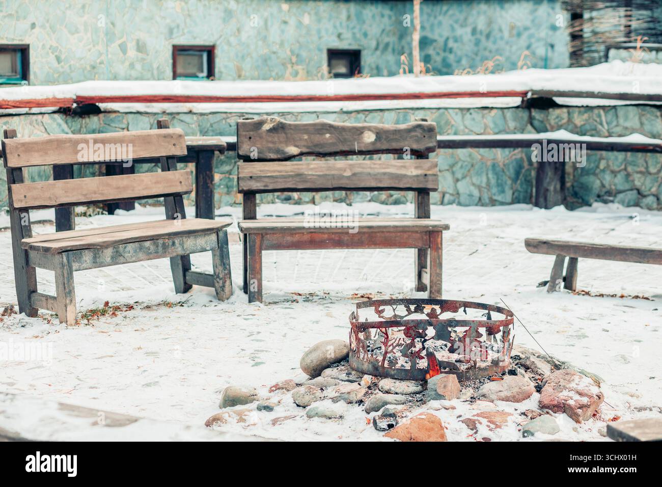 panche in legno intorno al focolare con pietre e ceneri nel giardino innevato, area ricreativa all'aperto rustica invernale concetto di campeggio, turismo e avventura Foto Stock