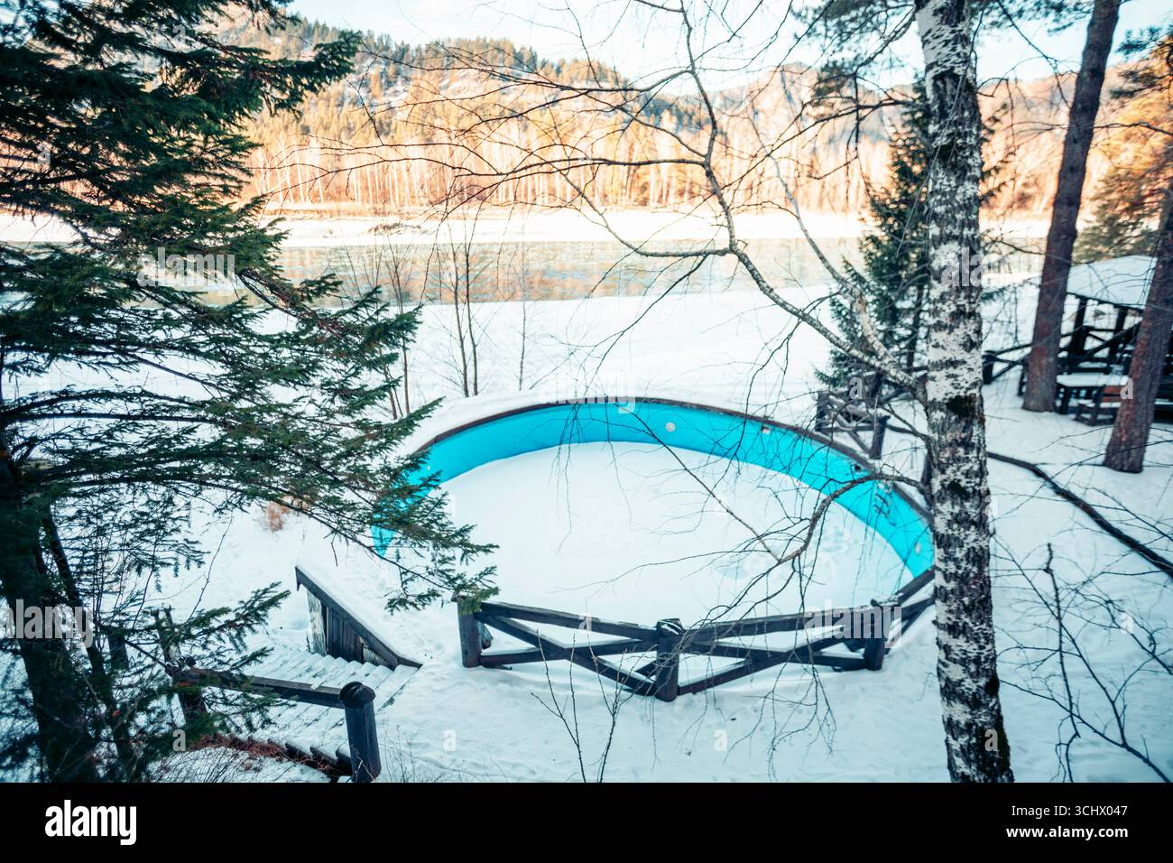 piscina all'aperto coperta di neve nella foresta invernale vicino al fiume e alle montagne, bacino blu vuoto circondato da recinzione in legno, turismo, stagionale recr Foto Stock