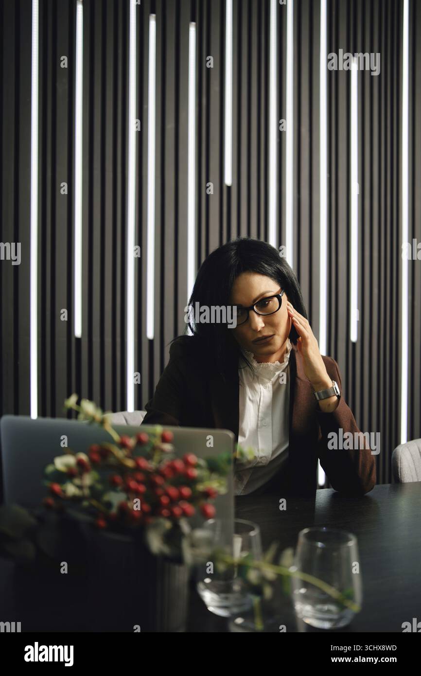 Una donna d'affari è seduta a una scrivania in un ufficio contemporaneo, con un'elegante illuminazione verticale che proietta ombre morbide. Lei è vestita Foto Stock