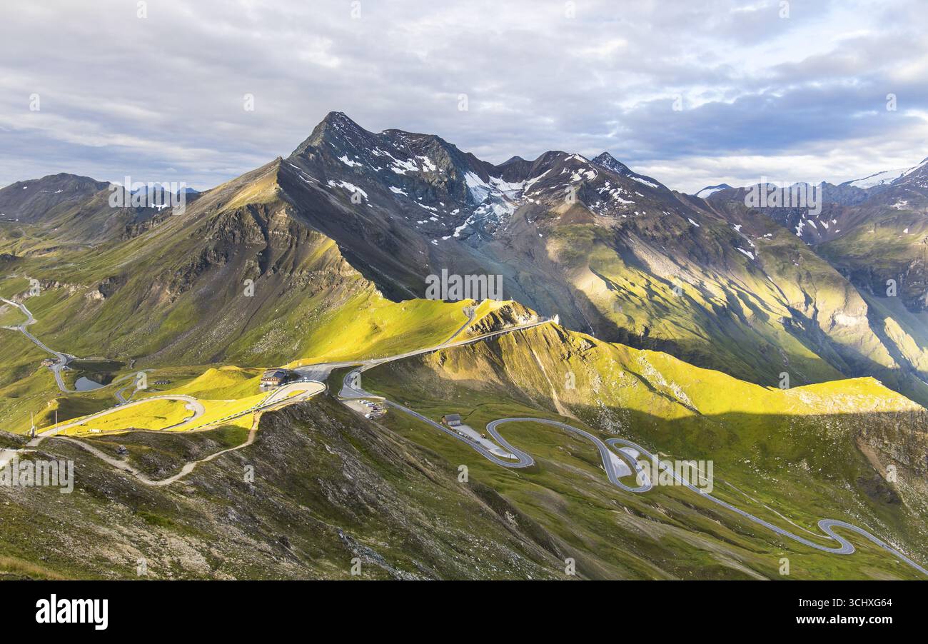 Veduta aerea delle strade tortuose che attraversano i pendii verdeggianti e baciati dal sole verso le aspre cime innevate della strada alpina Grossglockner, Bruck an der Großglocknerstraße, Salisburgo, Austria. Foto Stock