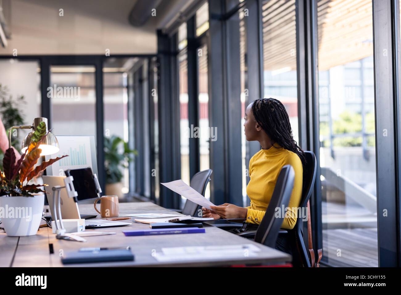 Donna d'affari in camicia gialla che esamina i documenti alla moderna scrivania dell'ufficio Foto Stock