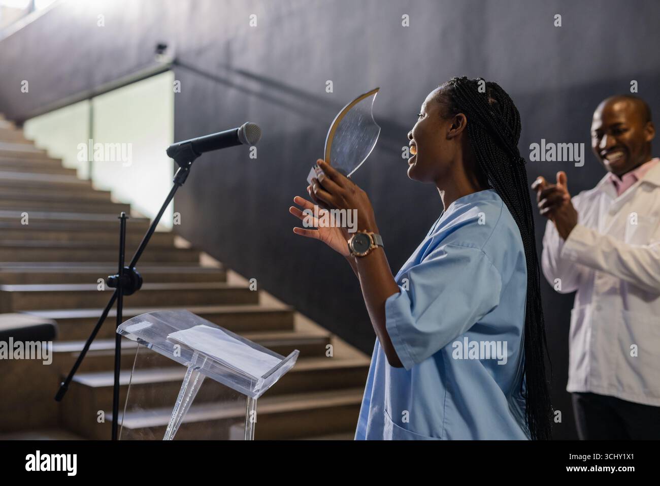 Donna afroamericana che tiene il premio, sorride e parla sul podio, uomo applaude Foto Stock