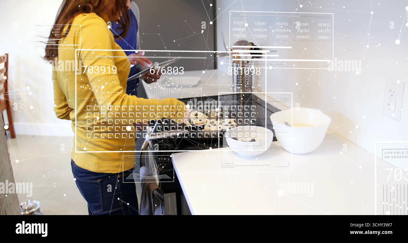 Cucinare una coppia che utilizza una stufa a gas in una cucina moderna, una donna che frigge il cibo e un uomo che legge un tablet Foto Stock