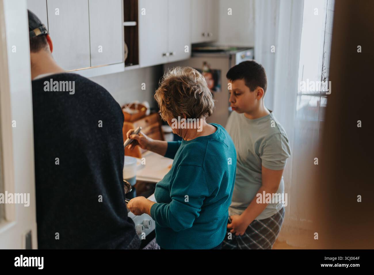 Donna anziana che cucina in una cucina di casa con i membri della famiglia che osservano e assistono con attenzione. Foto Stock
