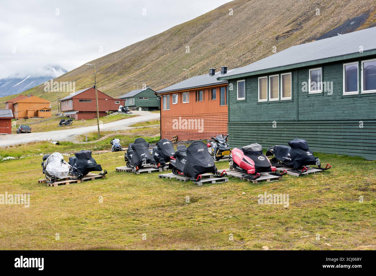 Le motoslitte Ski Doo Lynx Yamaha Longyearbyen Svalbard Norvegia // LONGYEARBYEN, SVALBARD - diverse motoslitte, tra cui i modelli Ski-doo Lynx e Yamaha, sono parcheggiate su pallet di legno in un'area erbosa di fronte agli edifici di Longyearbyen, Svalbard, Norvegia. I veicoli sono coperti da teloni protettivi, che suggeriscono di essere immagazzinati o in attesa di essere trasportati. Longyearbyen è il più grande insediamento e il centro amministrativo delle Svalbard, un arcipelago situato tra l'Oceano Artico e il Mare di Groenlandia. Il paesaggio presenta un ripido versante roccioso sullo sfondo, tipico dell'Artico Foto Stock