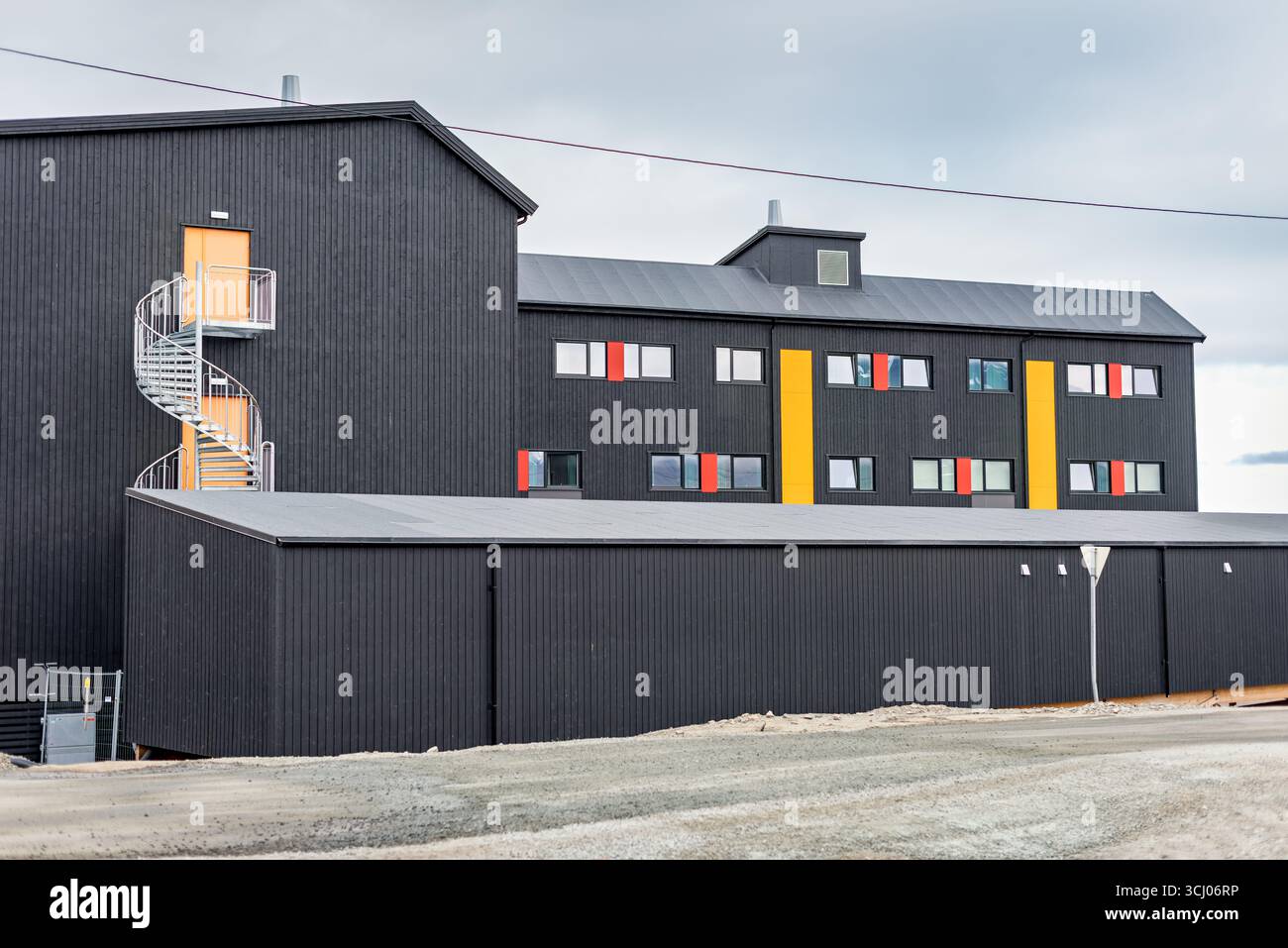 Architettura moderna con scalinata a spirale Longyearbyen Svalbard Norway // LONGYEARBYEN, SVALBARD - Un edificio architettonico moderno caratterizzato da una prominente scala a chiocciola che conduce ad una porta arancione contro una facciata di legno scuro. La struttura si trova a Longyearbyen, il più grande insediamento e centro amministrativo dell'arcipelago delle Svalbard, un territorio norvegese situato tra la Norvegia continentale e il Polo Nord. Le Svalbard sono note per la sua natura selvaggia artica, i ghiacciai e la popolazione di orsi polari, rendendo Longyearbyen un centro unico per la ricerca e il turismo in un ambiente aspro e remoto. Il bui Foto Stock