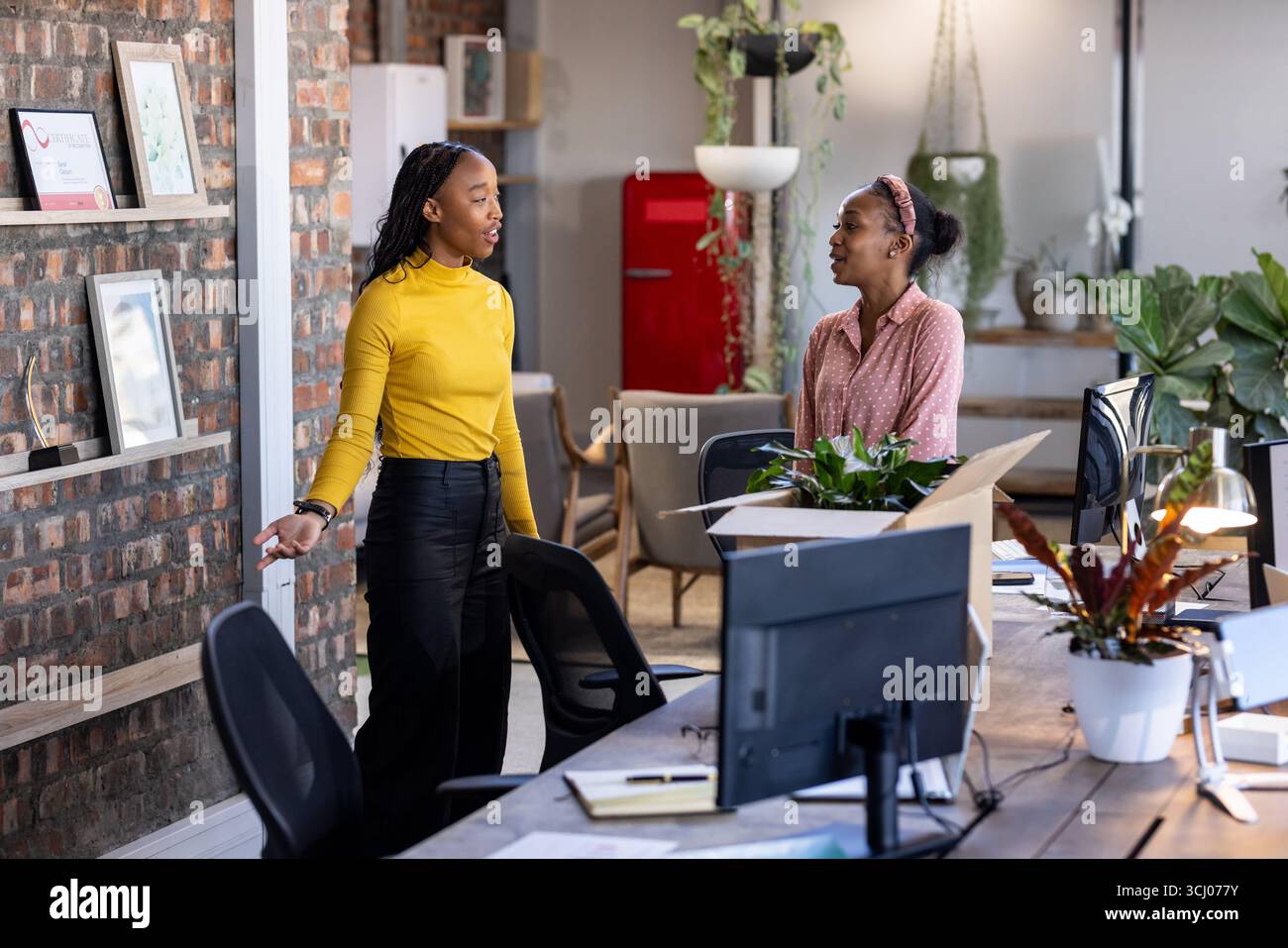 Due donne discutono di progetti in un ufficio moderno con piante e spazi di lavoro aperti Foto Stock