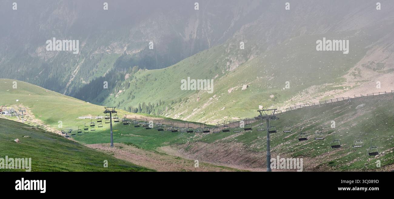 Vista panoramica di uno skilift vuoto che si estende su una valle di montagna verde nebbioso in estate. Paesaggio tranquillo con pendii erbosi, nebbia e lontano Foto Stock