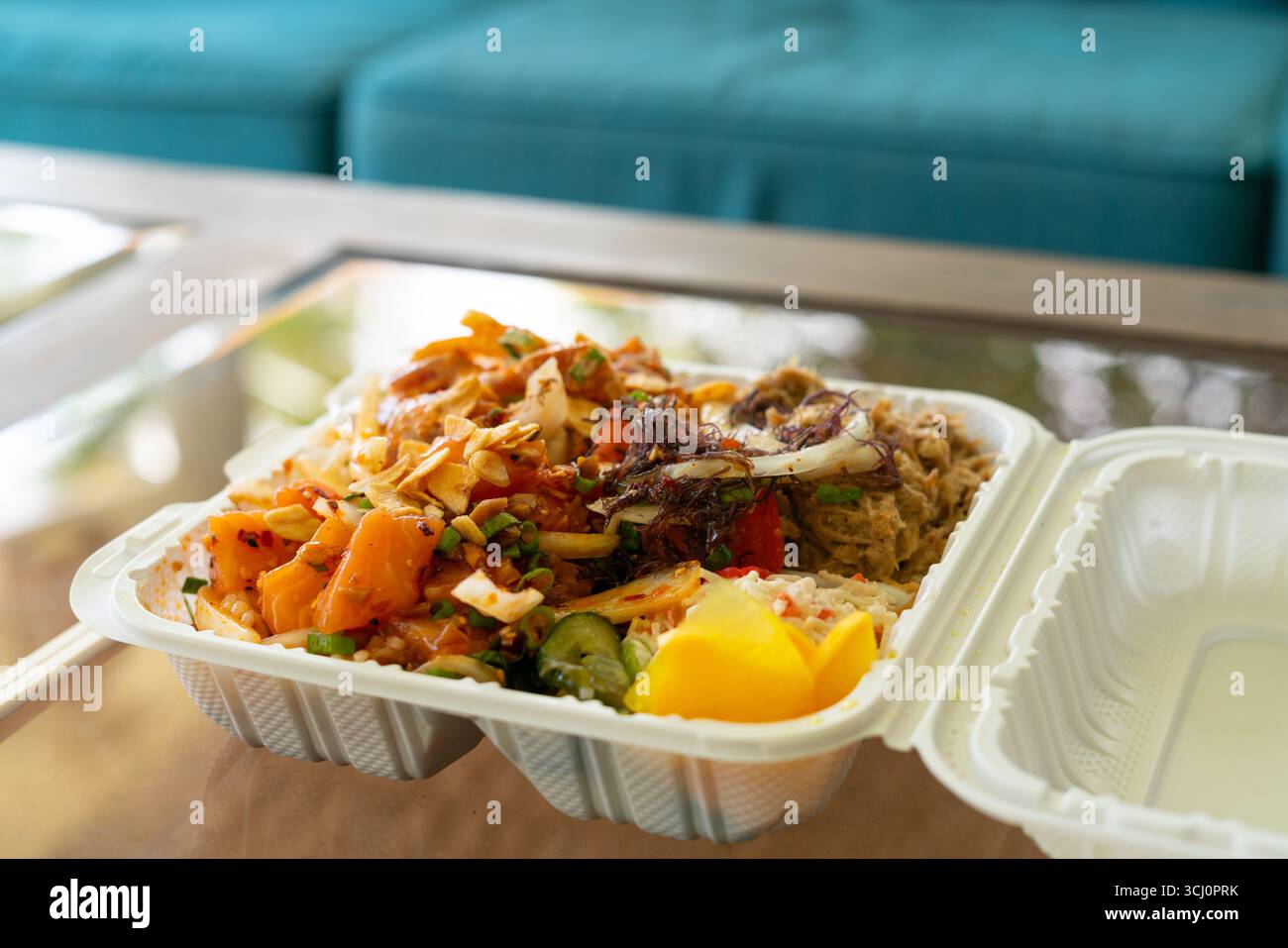 Colorato piatto hawaiano con salmone, ahi piccante, maiale di kalua e lati dell'isola, servito in una scatola da asporto su un tavolo di vetro, ideale per cene informali Foto Stock