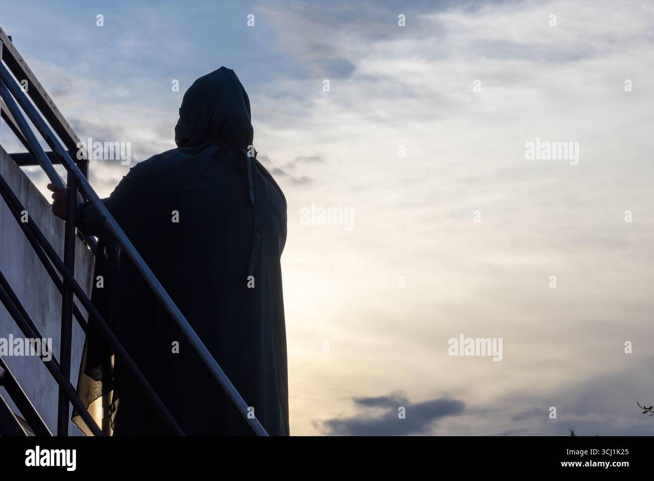 Silhouette di una donna musulmana bengalese premurosa in un abaya e un khimar, in piedi su un tetto e ammirando la vista mozzafiato del tramonto. Foto Stock