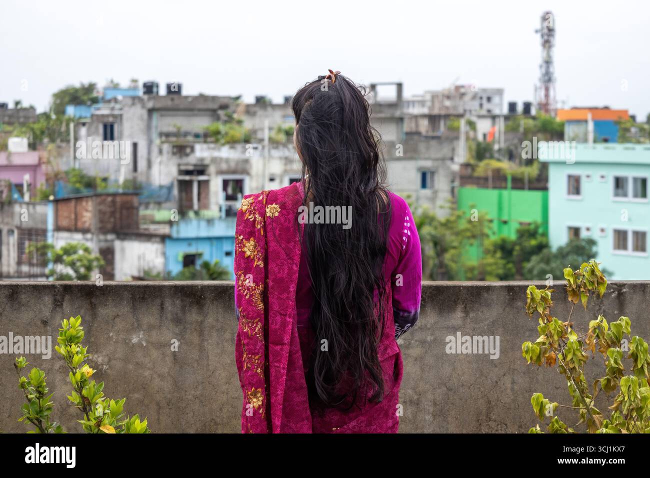 Vista posteriore di una splendida donna bengalese con lunghi capelli scuri, vestita con un sari di colore marrone, in piedi sul tetto della città e guardando gli edifici urbani in Foto Stock