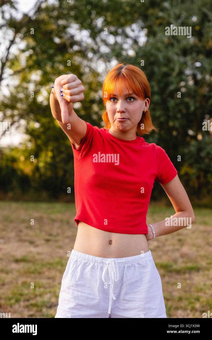 Una giovane donna che mostra un gesto con i pollici giù nel parco che esprime disapprovazione Foto Stock