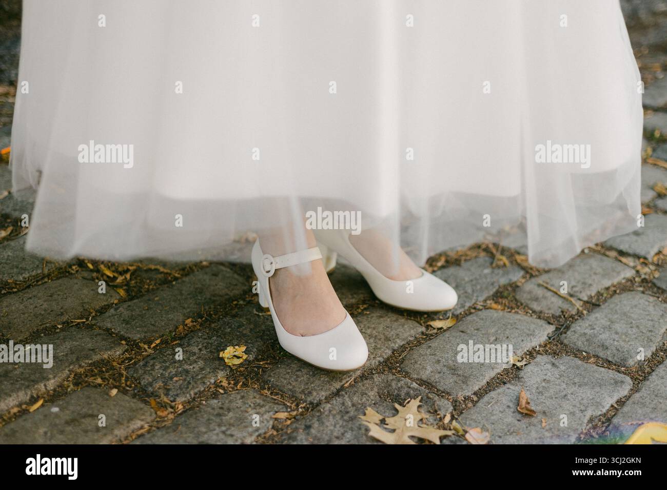 Le squisite scarpe bianche di una sposa sono parzialmente visibili sotto il suo abito fluente su un affascinante sentiero acciottolato. Foto Stock