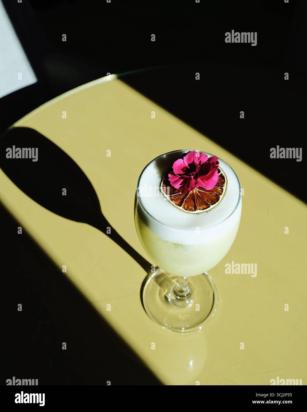 Un cocktail elegante e artigianale è presentato ad arte in un bicchiere a stelo con una testa spessa e spumeggiante. La bevanda è ricoperta da un fiore commestibile Foto Stock