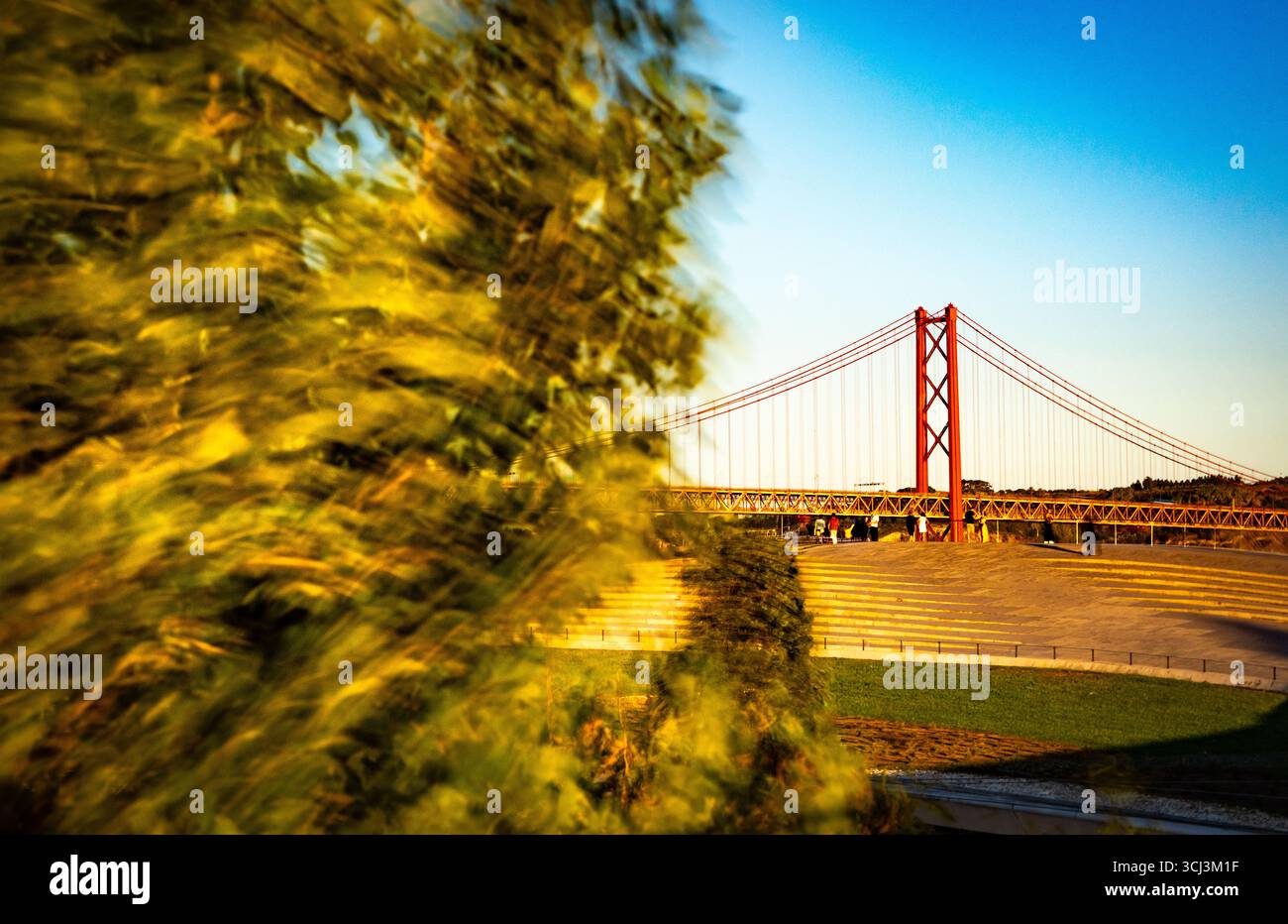 Il ponte 25 de Abril sul fiume Tago a Lisbona al tramonto con un albero in movimento in una giornata ventosa Foto Stock