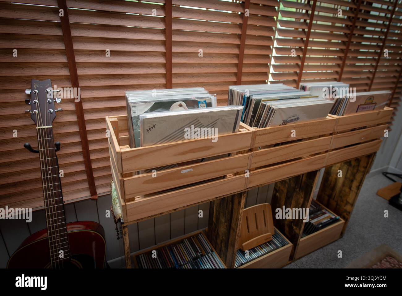 Un angolo di musica casalinga con casse di legno piene di dischi in vinile, CD sottostanti e una chitarra acustica nelle vicinanze, evocando un'atmosfera calda e nostalgica per la musica e. Foto Stock