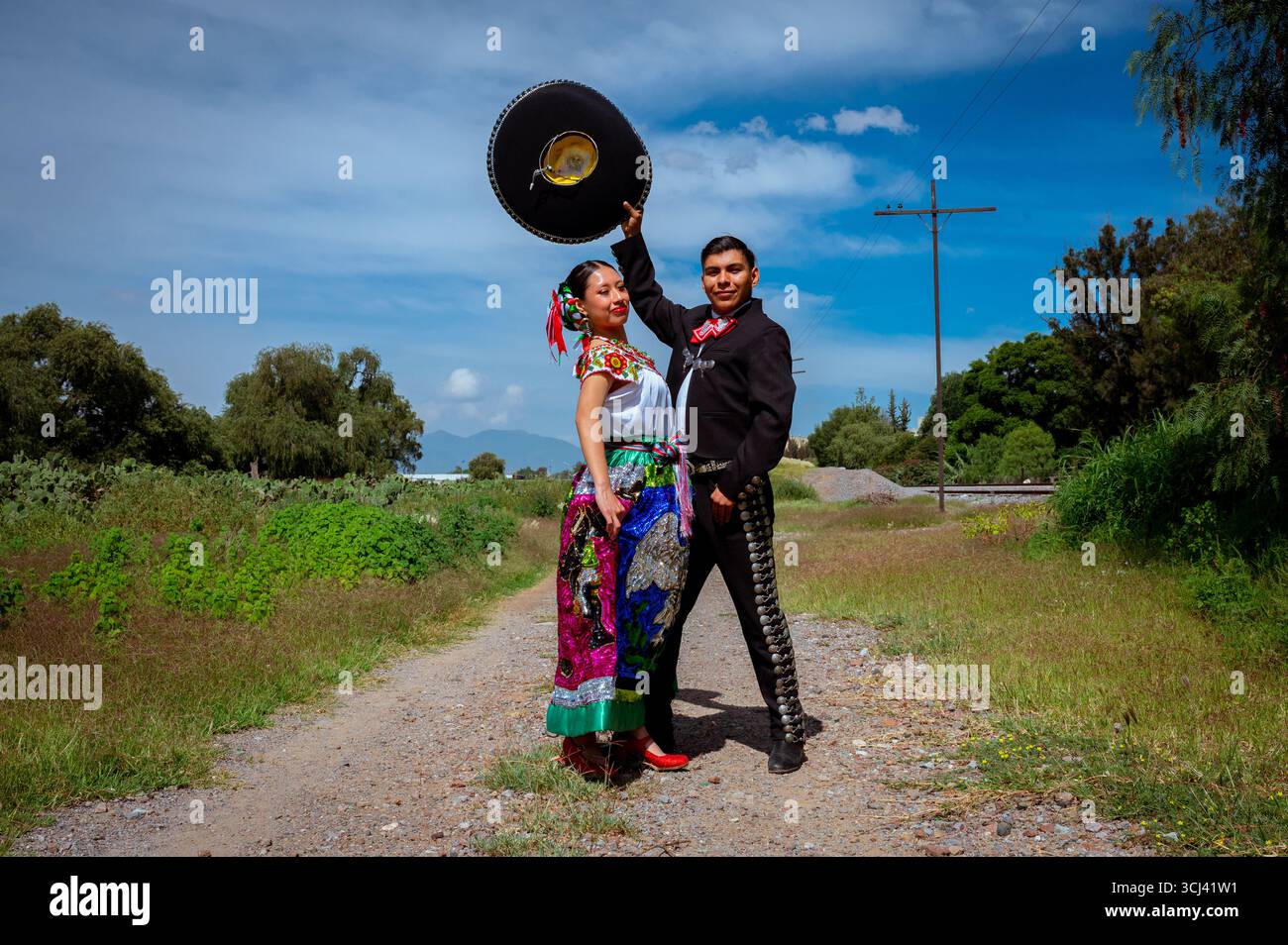 Coppia messicana che posa all'aperto in costumi folcloristici, con l'uomo che indossa un abito charro e un sombrero e la donna in un colorato abito ricamato Foto Stock