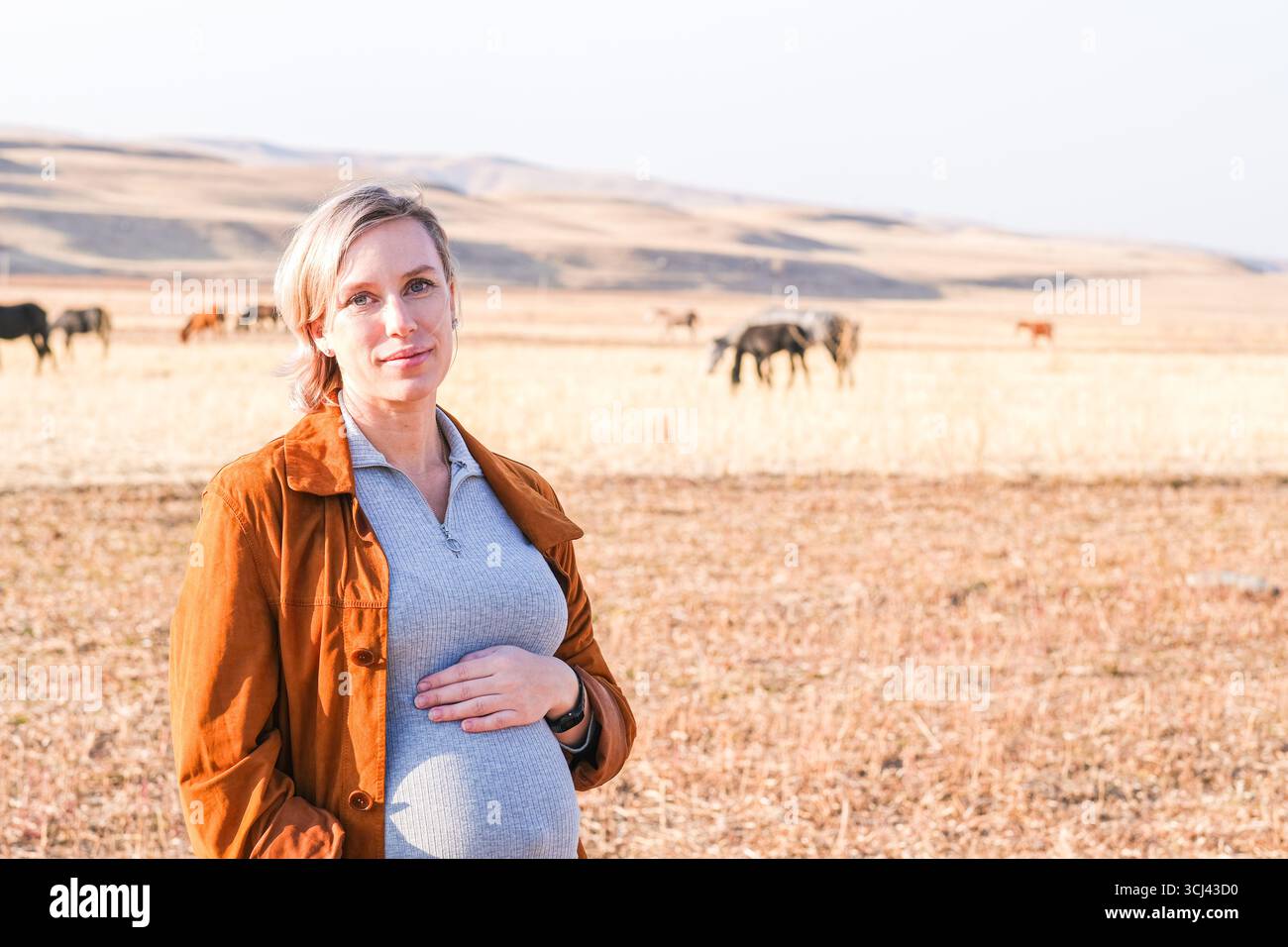 Donna incinta caucasica che tocca la pancia con cavalli che pascolano dietro. Viaggio di gravidanza nella natura. Esperienza di maternità. Foto Stock