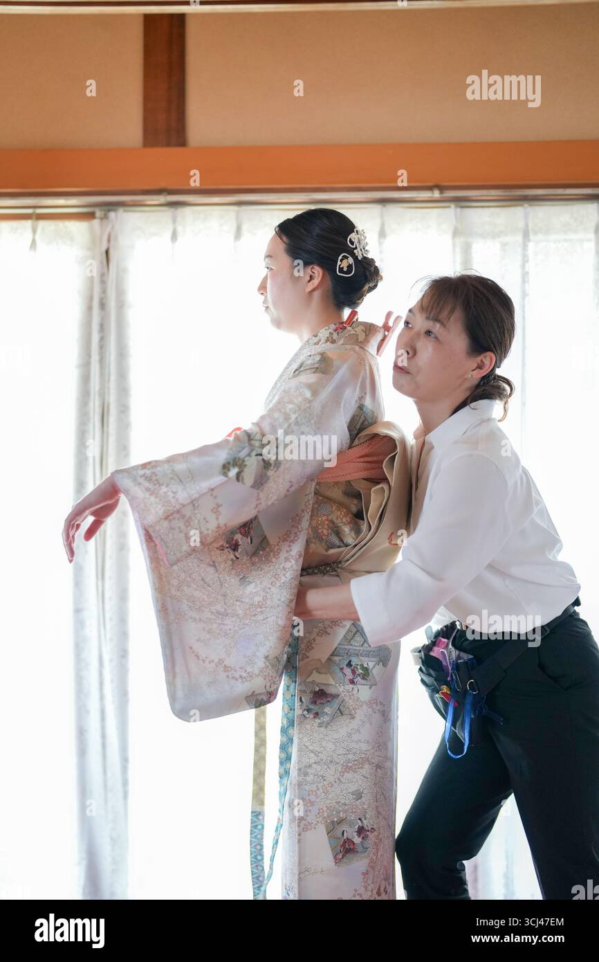 Una donna si sta facendo alterare il suo vestito da un'altra donna. L'abito è un tradizionale kimono giapponese. Le due donne sono in piedi in una stanza con una finestra Foto Stock